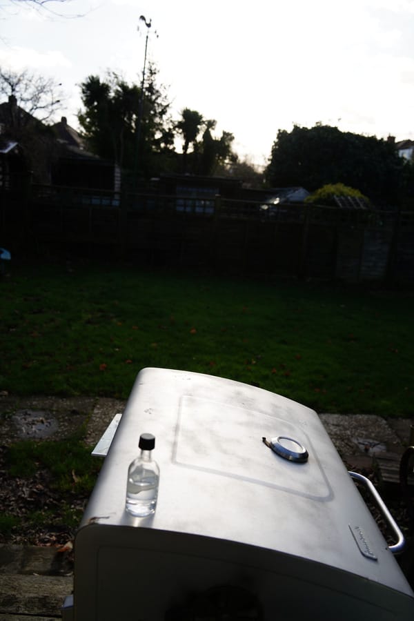 Small glass bottle placed on a silver barbecue lid in a garden, positioned to catch strong backlighting from the sun.